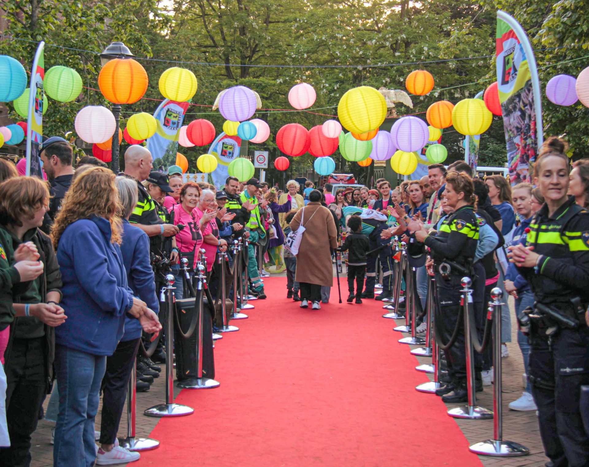 Vrouw met wandelstok loopt samen met haar kind over rode loper terwijl vrijwilligers klappen